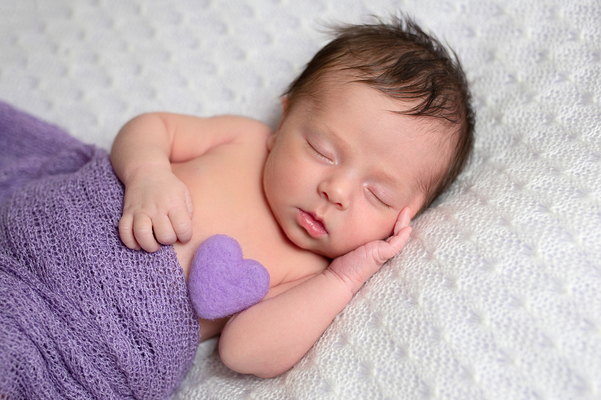 Nouveau-né endormi enveloppé dans un drap violet, tenant un petit cœur en laine